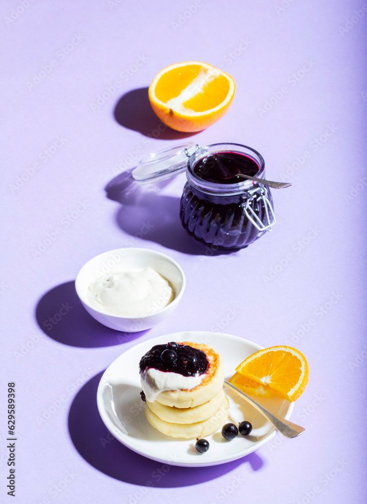 Breakfast of syrniki with sour cream and blackcurrant jam with orange on a lilac background.