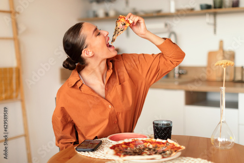 Young caucasian woman biting a slice of home cooked pizza, enjoying tasty dinner at home, sitting at table in kitchen