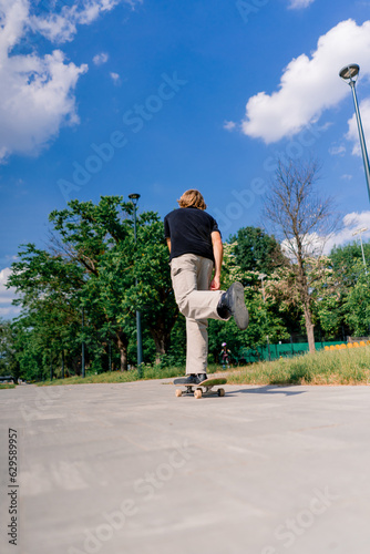 young guy skater rides a skateboard on the path of the city park against the background of trees and sky