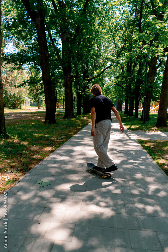 young guy skater rides a skateboard on the path of the city park against the background of trees and sky