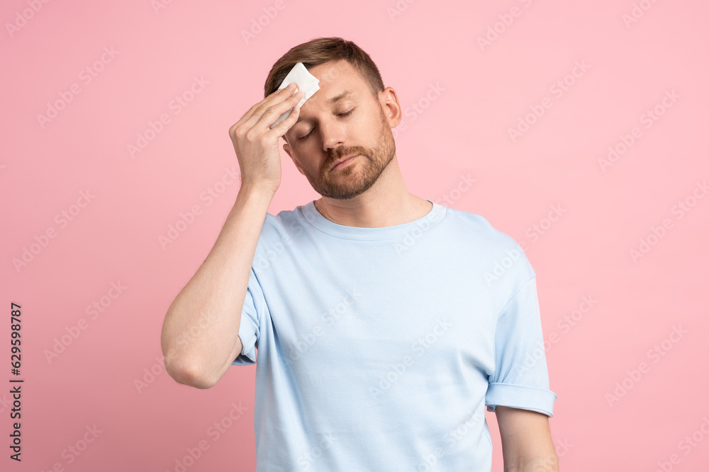 Man suffering from heat stuffiness wiping sweat on forehead with paper napkin isolated on pink background. Exhausted overheated bearded male with closed eyes. Summer hot weather, stuffy room concept. Man suffering from heat stuffiness wiping sweat on forehead with paper napkin isolated on pink background. Exhausted overheated bearded male with closed eyes. Summer hot weather, stuffy room concept.