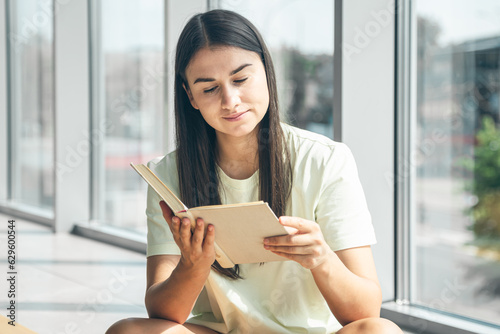 Attractive young woman reads a book while sitting by the window in summer.