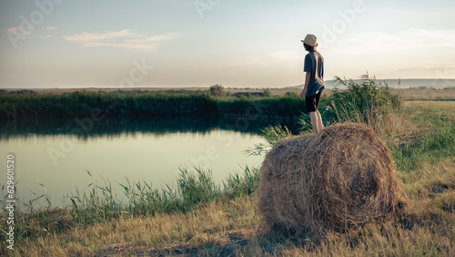 A young man in a hat stands on a haystack and looks at the surface of the lake in the reeds
