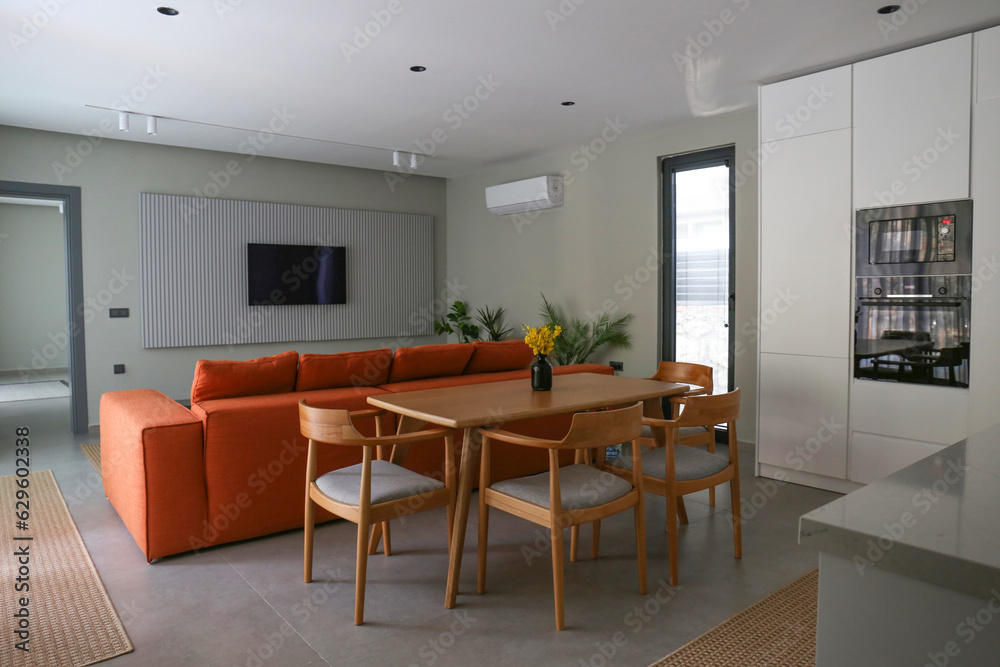 Open plan living room with orange textile couch and white cupboard kitchen with built-in appliances. Copy space, background. Open plan living room with orange textile couch and white cupboard kitchen with built-in appliances. Copy space, background.