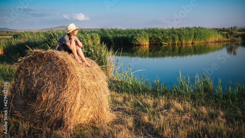 A young man in a hat sits on a haystack and looks at the surface of the lake in the reeds
