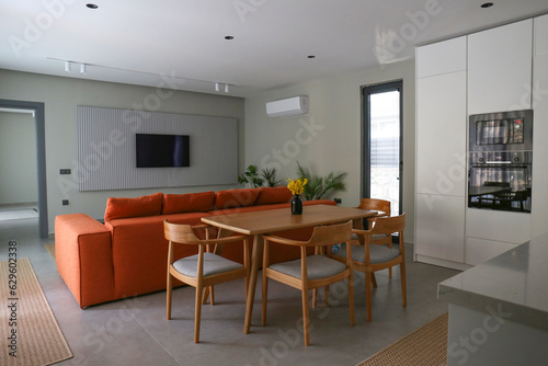Open plan living room with orange textile couch and white cupboard kitchen with built-in appliances. Copy space, background.