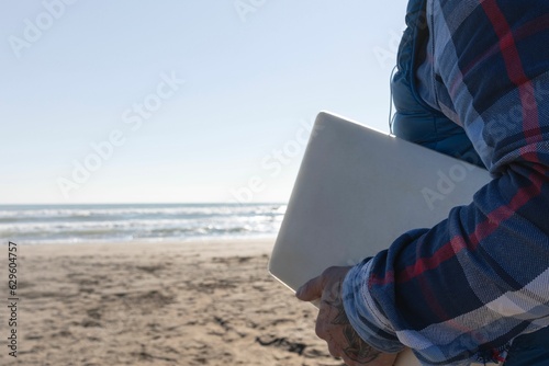 Young adult male standing on a beach with a laptop computer in his hand