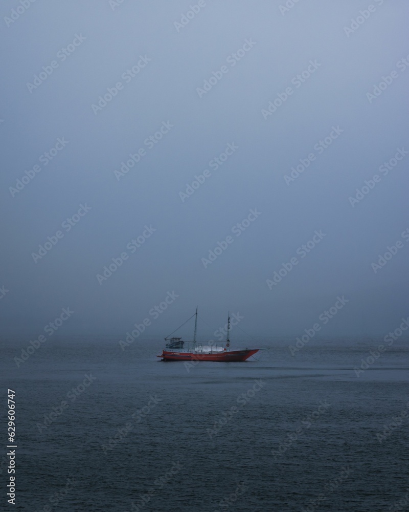 a red boat floating across the blue water in foggy day