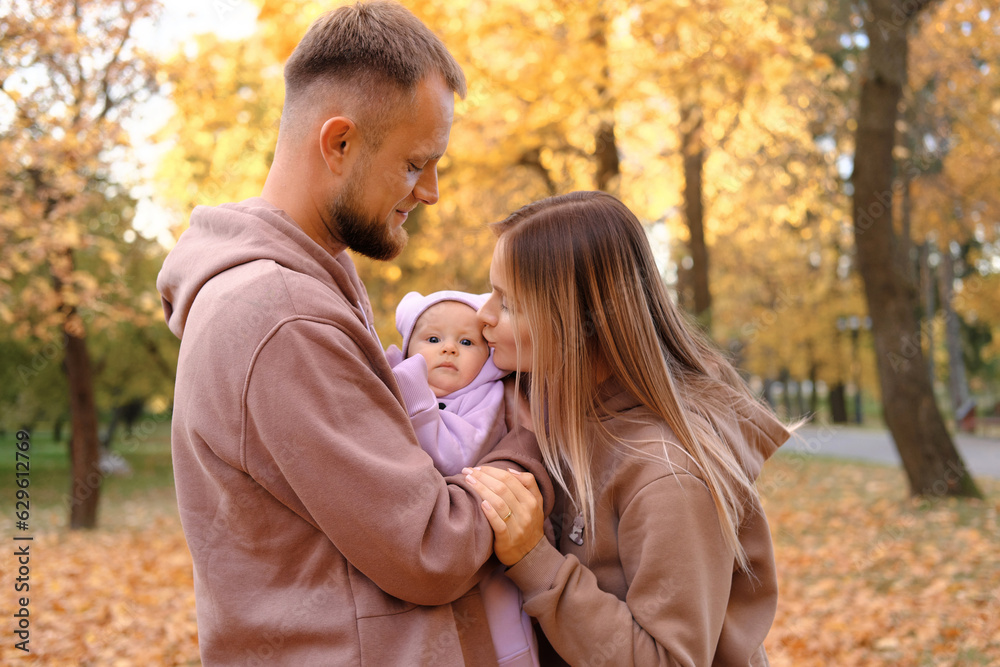 Young mother and father are walking with a newborn baby in the autumn park. Mother kissing newborn daughter Young mother and father are walking with a newborn baby in the autumn park. Mother kissing newborn daughter