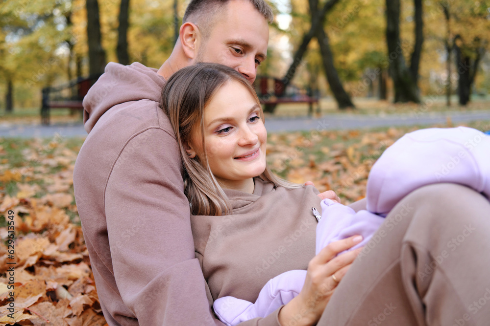 Naklejka premium Young parents sit on a plaid with a newborn baby in the autumn park. The child lies on the lap of the mother, the parents play with the child