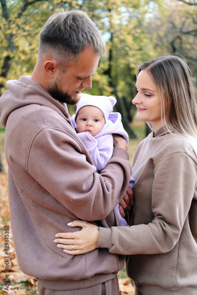 Fototapeta premium Young mother and father are walking with a newborn baby in the autumn park. Parents look at the child with tenderness