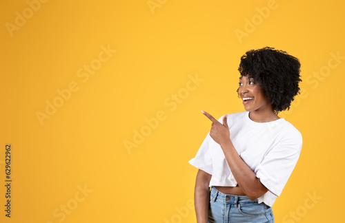 Cheerful surprised young black curly lady in white t-shirt pointing finger at copy space
