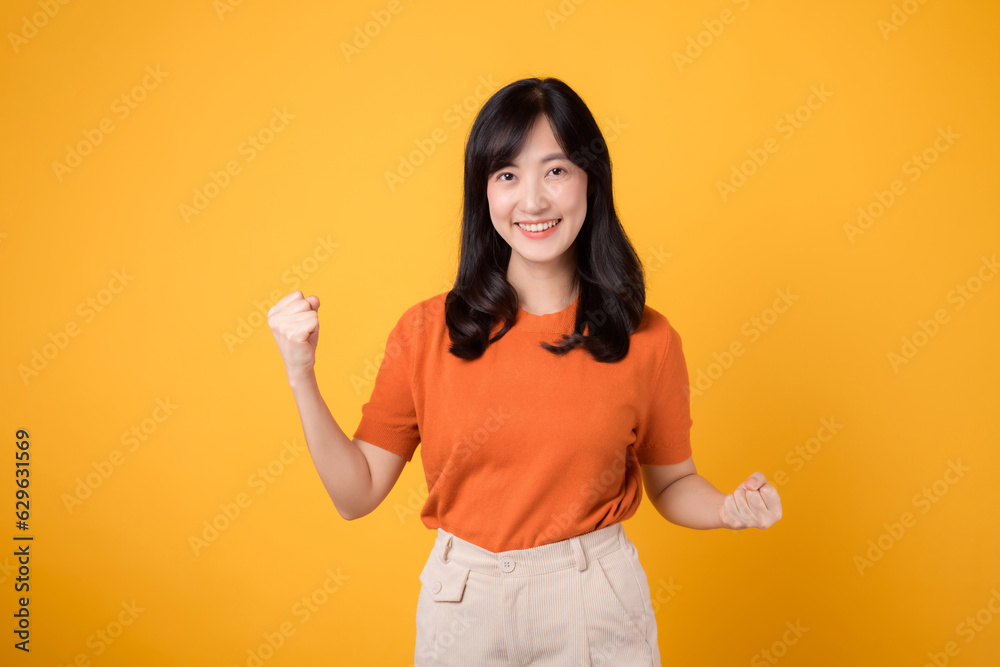 Embrace confidence as a young Asian woman in her 30s raises a fist up hand sign gesture, wearing an orange shirt on yellow background. Embrace confidence as a young Asian woman in her 30s raises a fist up hand sign gesture, wearing an orange shirt on yellow background.