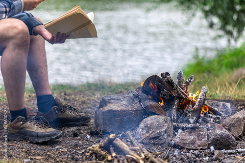 Close-up of bonfire with man reading a book in the background in nature.