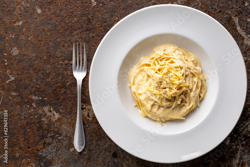 spaghetti carbonara with cream sauce in classy white circle ceramic plate beside fork on rusty texture background with copy space for text, top view, flat lay