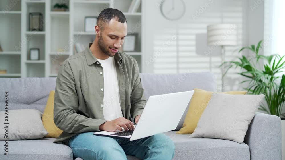 Young handsome african american man uses laptop computer while sitting on sofa at home. Mixed race male typing browsing web or social media shopping chatting working order online remotely indoor Young handsome african american man uses laptop computer while sitting on sofa at home. Mixed race male typing browsing web or social media shopping chatting working order online remotely indoor