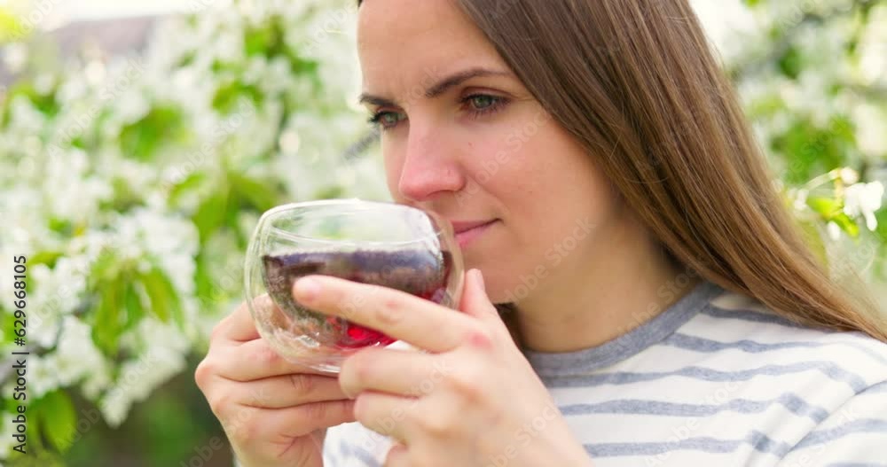 Young woman drinking tea in glass cup among the flowering trees in the garden Young woman drinking tea in glass cup among the flowering trees in the garden