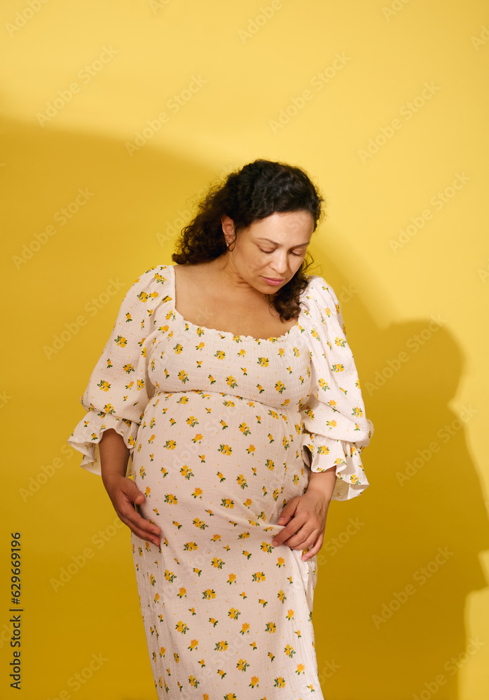Obraz premium Studio shot of a beautiful curly haired pregnant woman in summer sundress, isolated over yellow studio background
