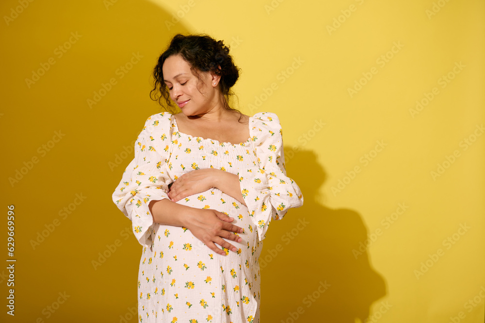 Delightful pregnant woman gently stroking and hugging her big belly, isolated over yellow studio background Delightful pregnant woman gently stroking and hugging her big belly, isolated over yellow studio background