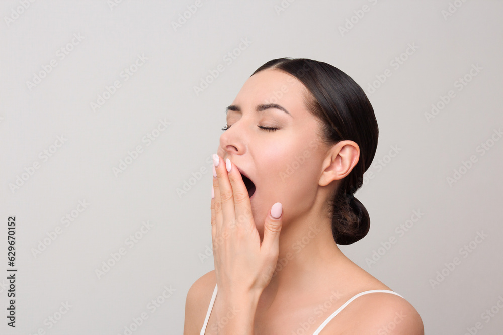 A young sleepy tired woman in white clothes yawns, covering her mouth with her hand, closing her eyes, isolated on a gray background. The concept of drowsiness and fatigue. Human emotion A young sleepy tired woman in white clothes yawns, covering her mouth with her hand, closing her eyes, isolated on a gray background. The concept of drowsiness and fatigue. Human emotion