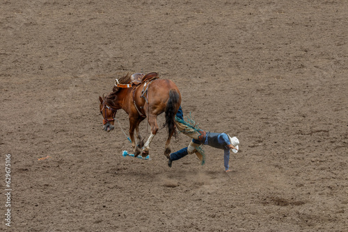 A cowboy is falling off a bucking bronco at a rodeo in an arena. The horse has all four legs off the ground. The cowboy is wearing blue with a white hat. They are in a dirt arena.