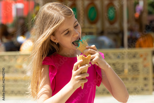 Adorable little girl in casual clothes eating delicious sweet ice cream while having fun on blurred background of amusement park