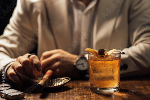 Man smoking a cigar and having an old-fashioned cocktail served on a wooden bar table