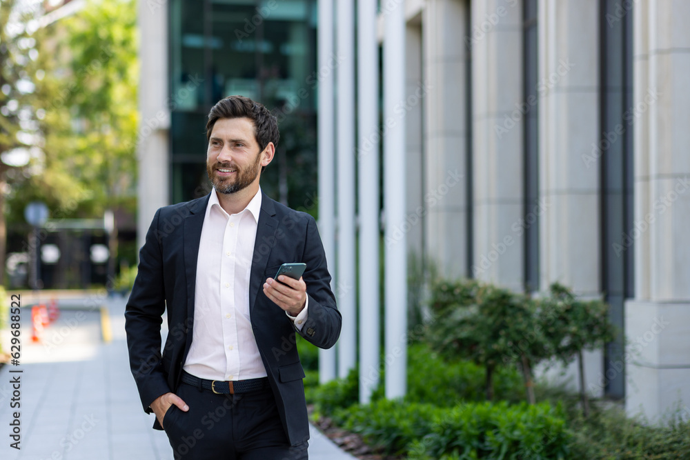 Smiling young businessman man in a business suit is using a mobile phone on the street, walking on a break outside, waiting for a meeting Smiling young businessman man in a business suit is using a mobile phone on the street, walking on a break outside, waiting for a meeting
