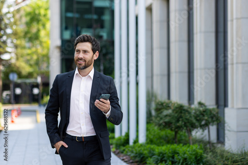 Smiling young businessman man in a business suit is using a mobile phone on the street, walking on a break outside, waiting for a meeting