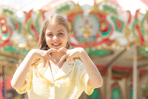 Kindness Concept. Portrait of cute blonde hair girl making heart shape with her hands and fingers, smiling to camera, posing while walking outdoor in a amusement park, copy space