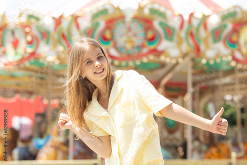 Adorable student blonde girl in summer yellow clothes at amusement park. Cute girl stands on the background of the carousel and laughs. Entertainment concept