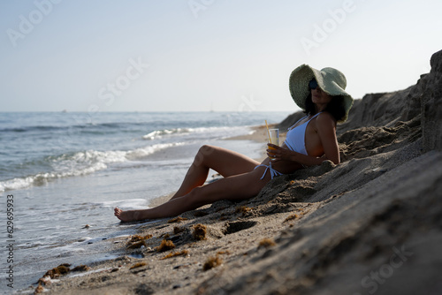 beautiful tanned brunette girl in a white bathing suit drinks from a glass of orange juice sitting on the sand on the beach by the sea
