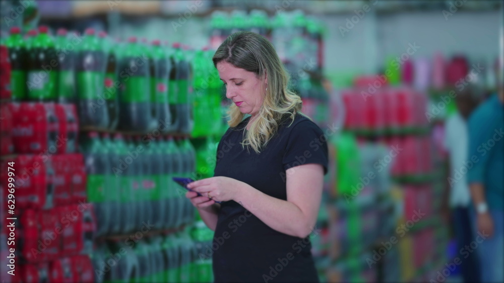 Female consumer walking through supermarket soda drinks aisle holding cellphone browsing products and looking at grocery list. Woman shopping depicting consumerism lifestyle habits