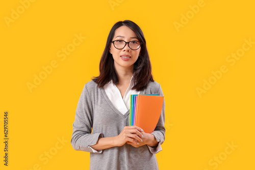 Female Asian teacher with copybooks on yellow background