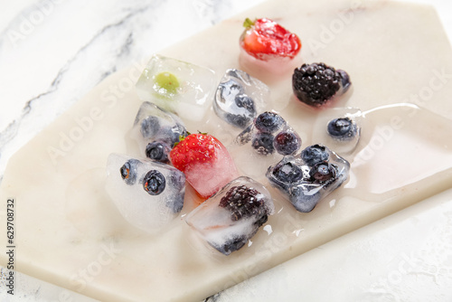 Board with frozen berries in ice cubes on white background