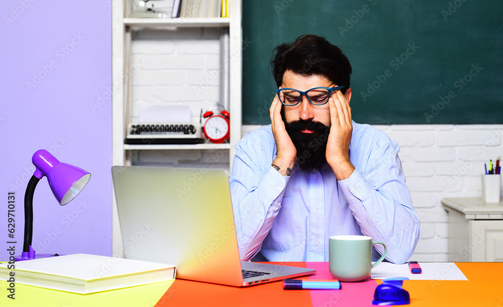 Exhausted stressed teacher suffering from headache sitting at desk in classroom. Tired male student feeling migraine after huge load of computer working. University professor thinking about problems. Exhausted stressed teacher suffering from headache sitting at desk in classroom. Tired male student feeling migraine after huge load of computer working. University professor thinking about problems.