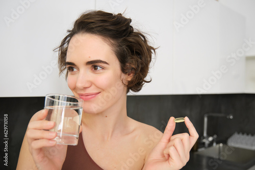 Portrait of sportswoman drinking water, taking vitamins, dietry supplements for healthy skin, having omega-3 pill, standing in her kitchen in workout clothing photo