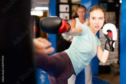 Determined athletic young woman in boxing gloves practicing heel kick by straight leg on heavy punching bag during workout in gym