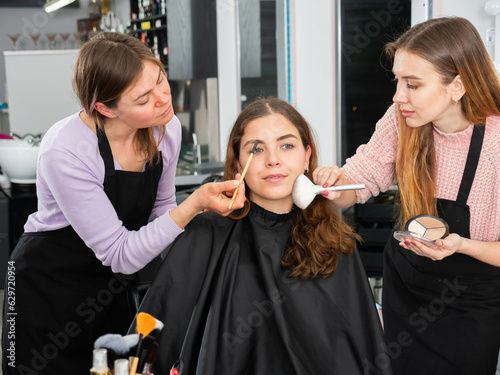 Two professional female makeup artists working in a beauty salon apply makeup on the face of a young woman photo