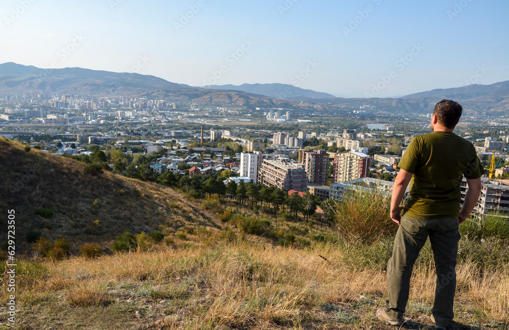 Back view of man standing on the hill and looking to the residential buildings and mountains in background at Tbilisi, Georgia