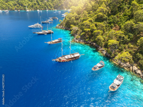 Aerial view of beautiful yachts and boats on the sea at in summer sunny day. Gemiler Island in Turkey. Top view of luxury yachts, sailboats, clear blue water, beach, mountain and green forest. Travel photo