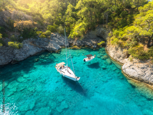 Aerial view of beautiful yacht and sailboat on the sea at sunrise in summer. Oludeniz lagoons, Turkey. Top view of bay, luxury yacht, motorboat, clear blue water, beach, rocks and green trees. Travel