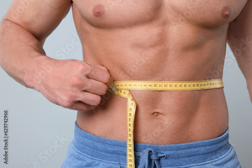 Athletic man measuring waist with tape on light grey background, closeup. Weight loss concept