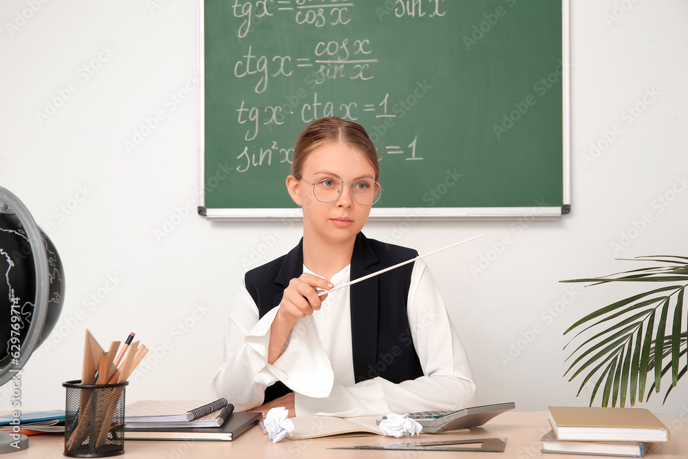 Young math teacher with pointer sitting at wooden table in classroom Young math teacher with pointer sitting at wooden table in classroom
