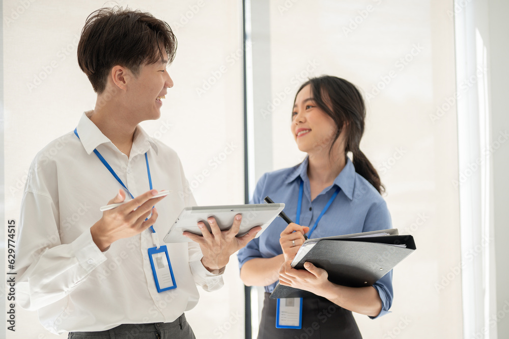 Two happy young Asian office workers are talking while standing in a company corridor together. Two happy young Asian office workers are talking while standing in a company corridor together.