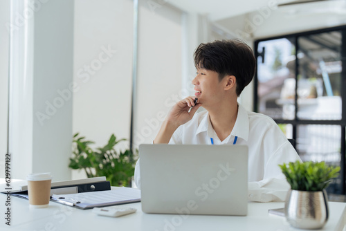 young businessman in white shirt sitting at desk holding pen and working on laptop. employee working at desk