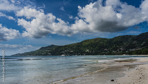 Beautiful tropical beach. The waves of the turquoise ocean spread over the sand. The yacht is far away. A green hill against a background of blue sky and clouds. Seychelles. Mahe. Beau Vallon.