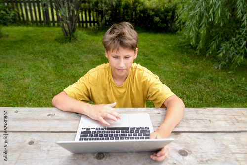 TTeen boy with a laptop sits in a park. School boy is typing on computer outdoors. Young boy studying online with a computer. Boy is online lesson while outside. photo