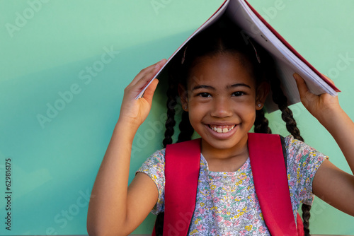Portrait of happy biracial schoolgirl with book on head over blue background at elementary school