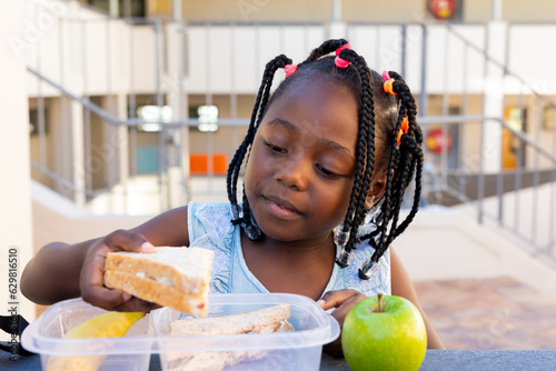 African american schoolgirl at table and having healthy lunch eating sandwich at elementary school